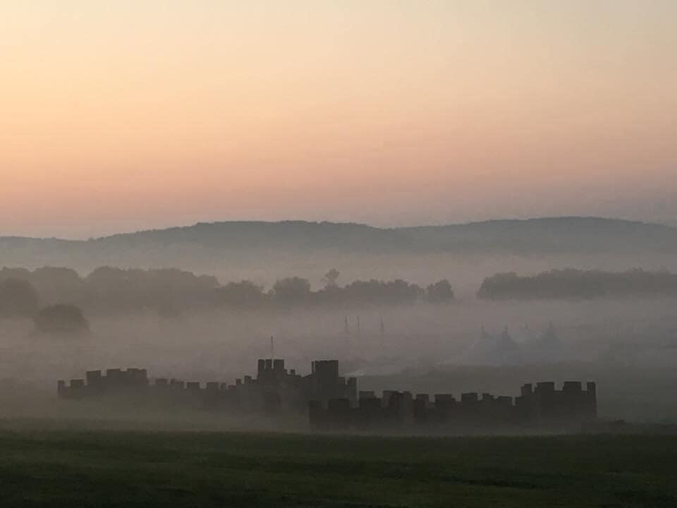 A picture of a rolling field with a wooden castle front below. Fog covers the low lying field with the tops of the trees emerging from it.